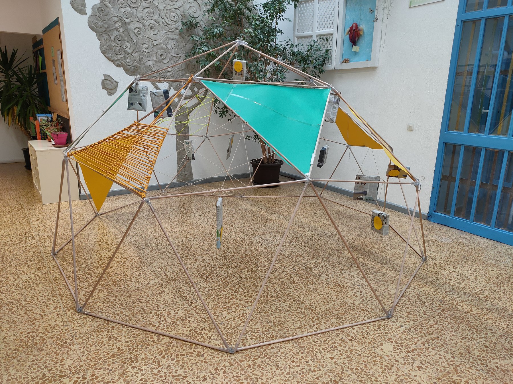 Estructura geodésica con superficies verdes y amarillas, cuerdas y objetos suspendidos en un patio interior.
