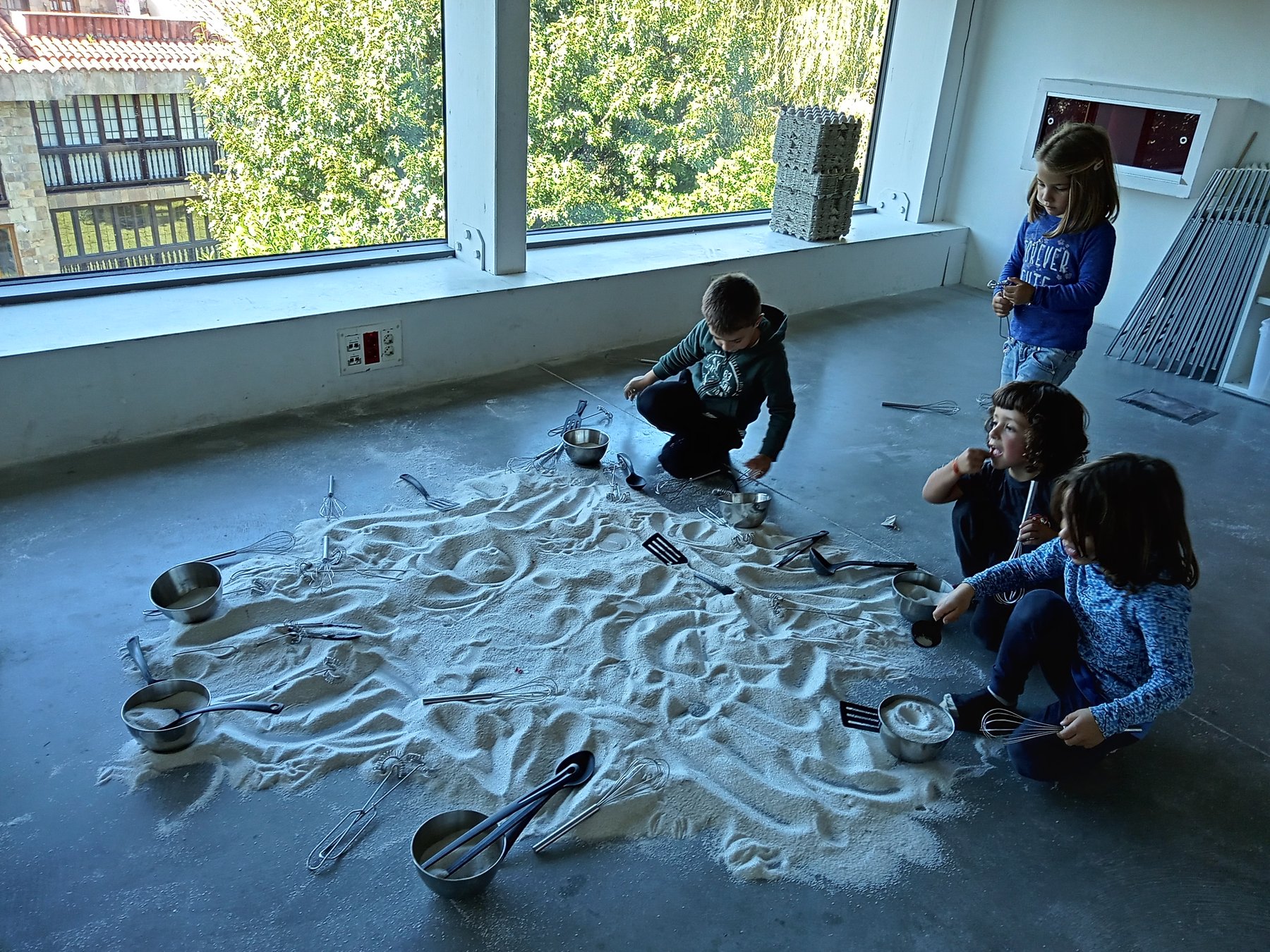 Niñas y niños exploran arena, utensilios metálicos y marcas en el suelo junto a una ventana.
