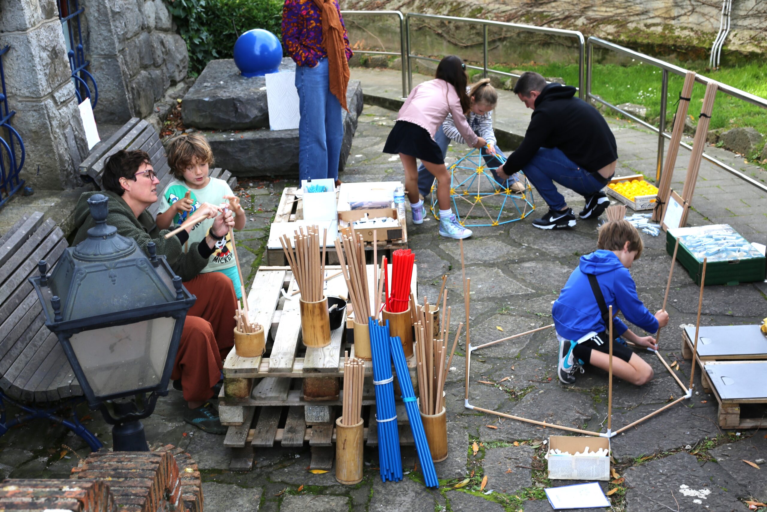 Personas adultas y niñas y niños exploran materiales y construcciones en un patio junto a un edificio de piedra.